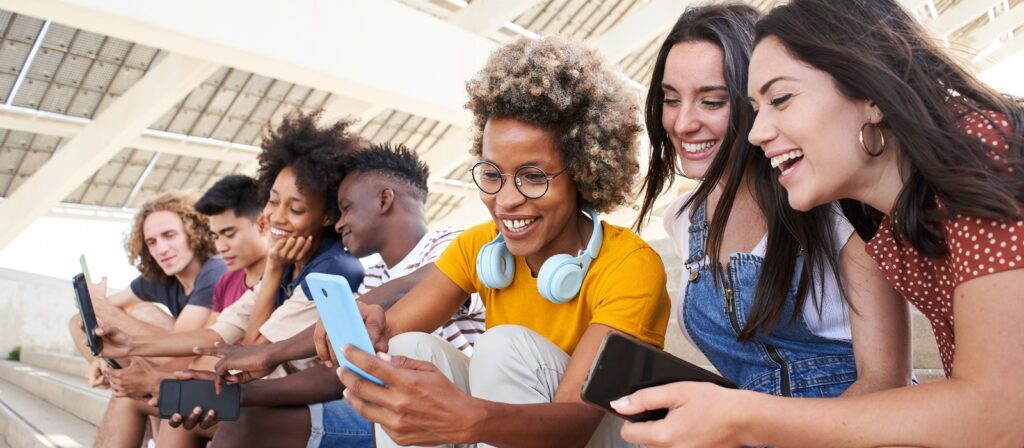 A group of seven smiling young adults sitting outdoors on steps, looking at mobile phones and laughing together. One person has headphones round their neck. It is a bright, sunny day.