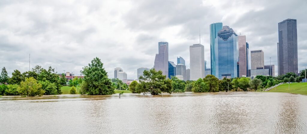 Floodwaters cover a park area with trees partially submerged in the foreground, as the skyline of downtown Houston, Texas, rises in the background under a cloudy sky.
