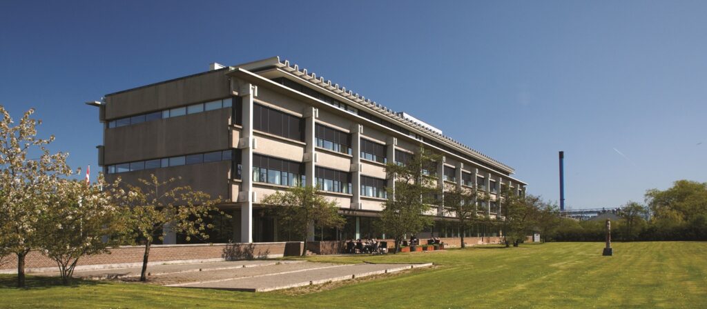 A modern, three-storey building with large windows sits beside a green lawn and scattered trees under a clear blue sky. People are seated at tables outside near the building.