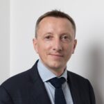 A man in a dark suit, light blue striped shirt, and navy tie stands against a white background, looking at the camera with a slight smile.