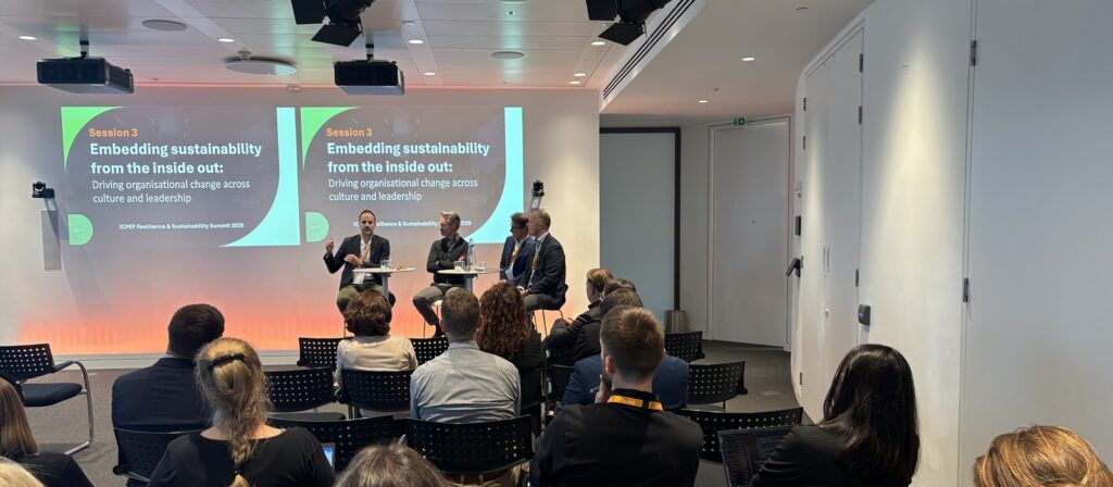 A panel of three speakers sits on a stage in front of an audience, discussing sustainability at a business event. Two large screens behind them display the session title: Embedding sustainability from the inside out.