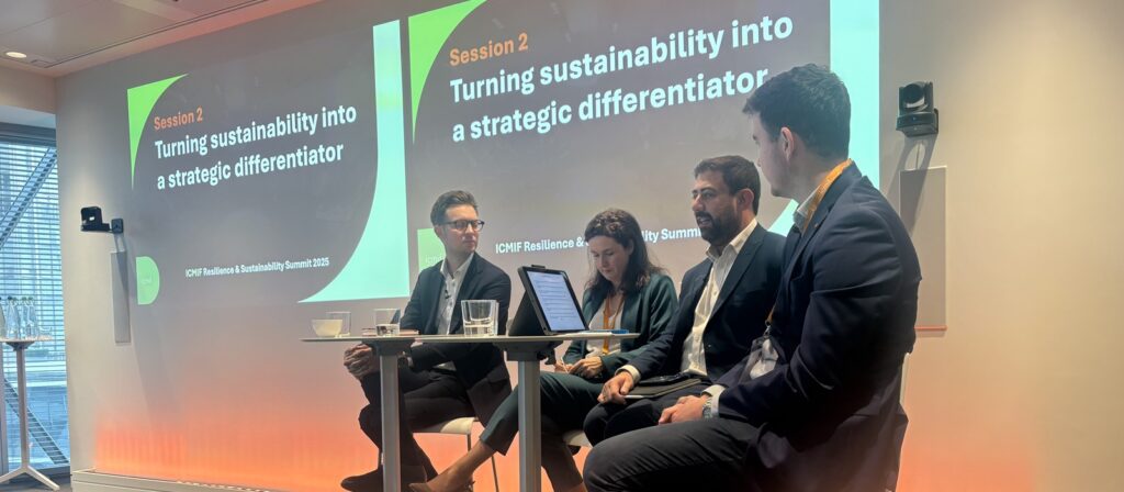 Four people sit on a panel in front of a projected screen at a conference. The screen displays Session 2: Turning sustainability into a strategic differentiator. Two men and a woman have laptops and notes.