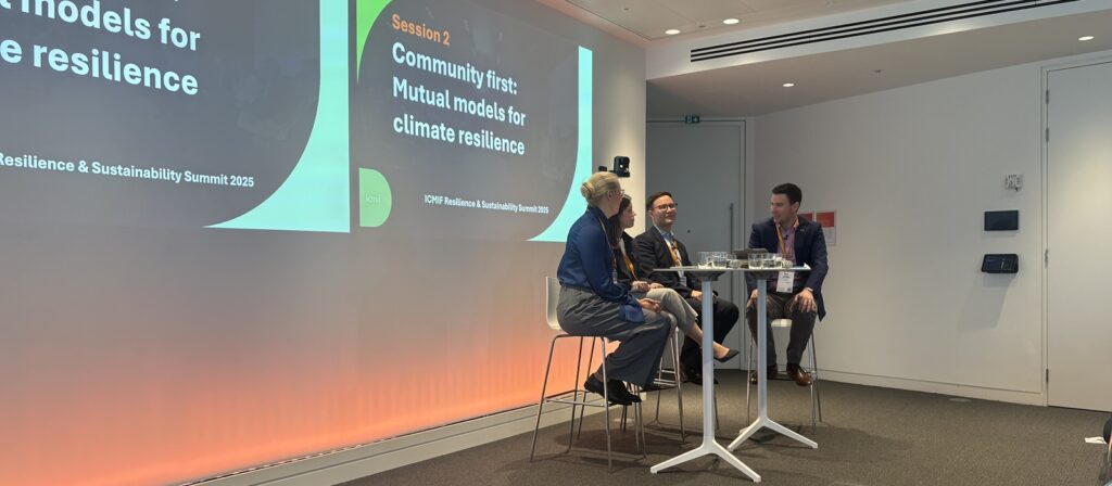 Three people sit on a stage in discussion during a conference session titled Community first: Mutual models for climate resilience. A large screen displays the session title. The setting is a modern, well-lit conference room.