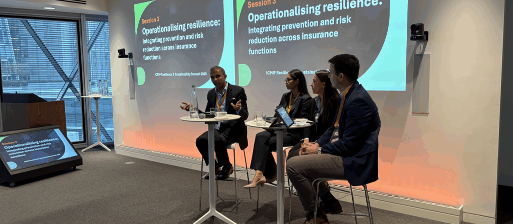 Four professionals sit on a stage panel, speaking and listening, with two screens behind them displaying the session title: Operationalising resilience: Integrating prevention and risk reduction across insurance functions.