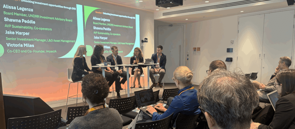 A panel of five speakers sits at the front of a modern conference room, addressing an audience. Two large screens behind them display the panellists' names and titles. Attendees sit in rows, some taking notes.