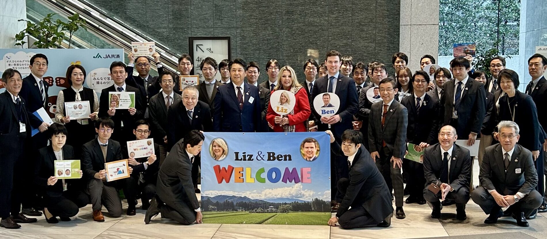 A large group of people in suits stand together indoors, holding signs and a colourful “Welcome Liz & Ben” banner with a scenic background. Two people in the centre hold round signs with their names and photos.