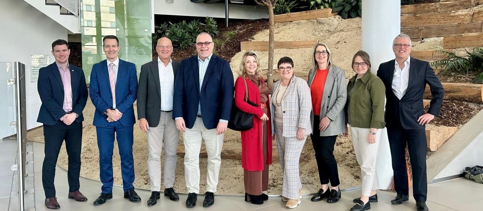 Nine people stand in a row indoors, smiling at the camera. They are dressed in business or smart-casual attire, with plants and landscaped features in the background.