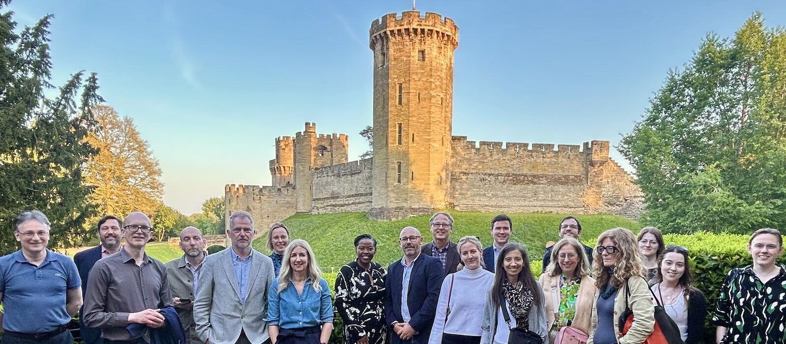 A group of people pose and smile for a photo outdoors in front of a historic stone castle with a tall round tower, surrounded by green trees and grass under a clear blue sky.