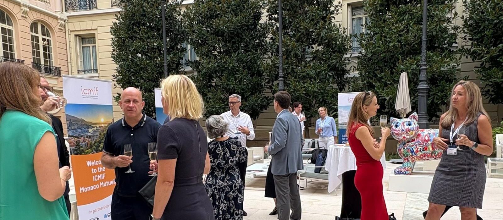 A group of people dressed in business attire converse and hold drinks at an outdoor networking event, with banners, tables, and a colourful cat sculpture visible in the background.