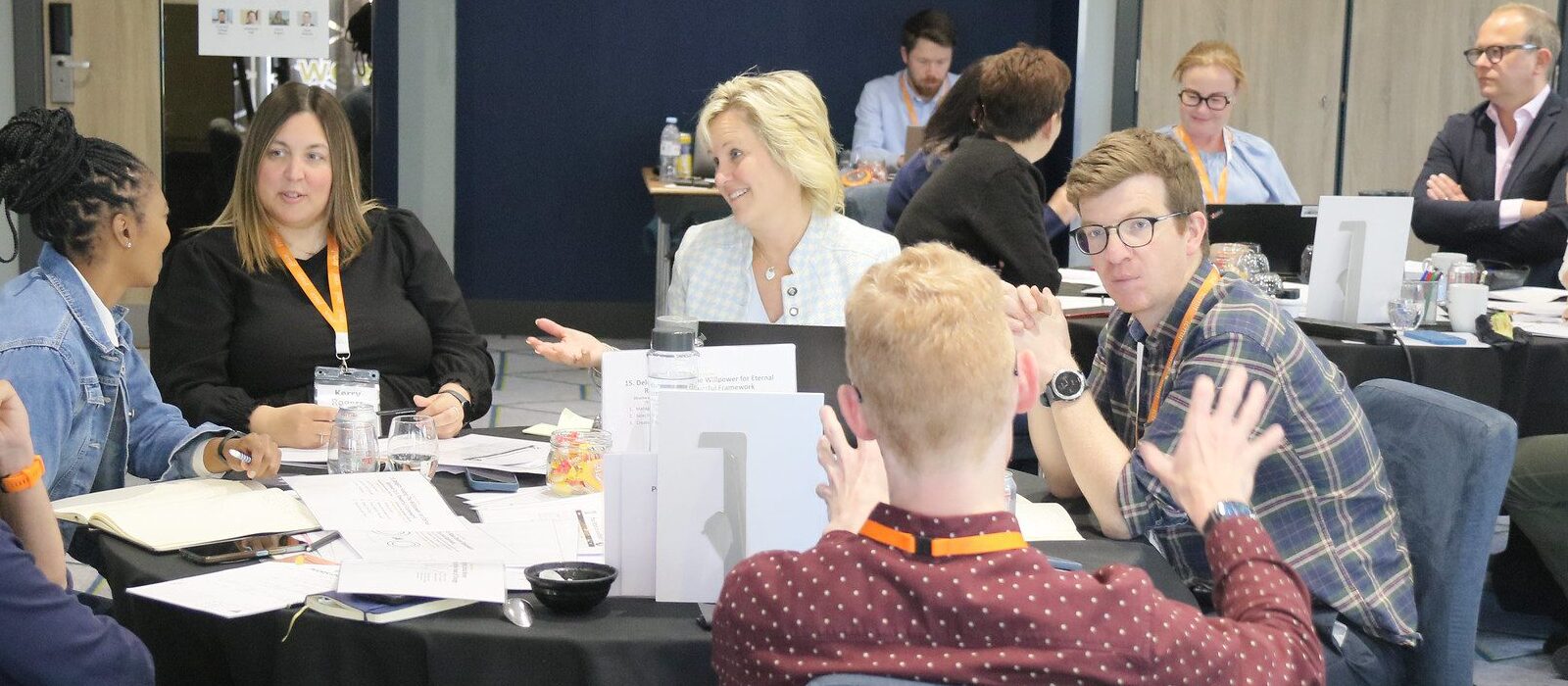 A group of people sit around a table, engaged in discussion at a workshop or meeting. Papers, notebooks, and drinks are on the table, and most participants are wearing lanyards. Other groups are visible in the background.