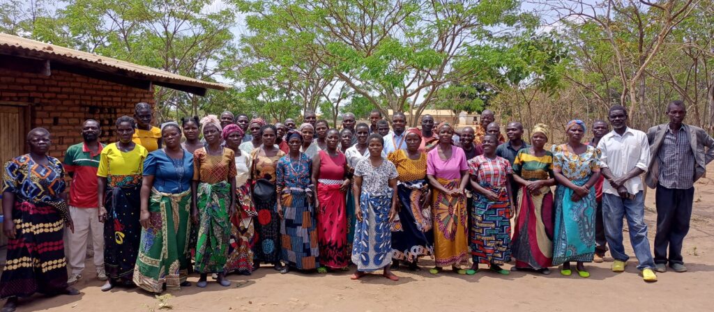 A large group of men and women stand outside in front of trees and buildings, smiling and dressed in colourful, patterned clothing. The setting appears to be in a rural area with clear skies.
