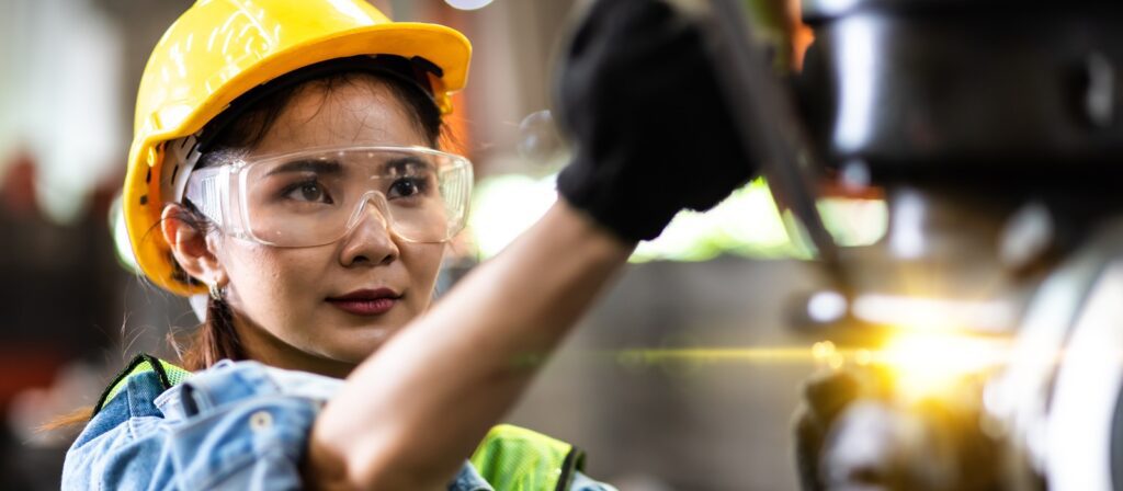 A woman wearing a yellow hard hat, safety glasses, and gloves operates machinery in an industrial setting, focusing intently on her task with visible sparks in the background.