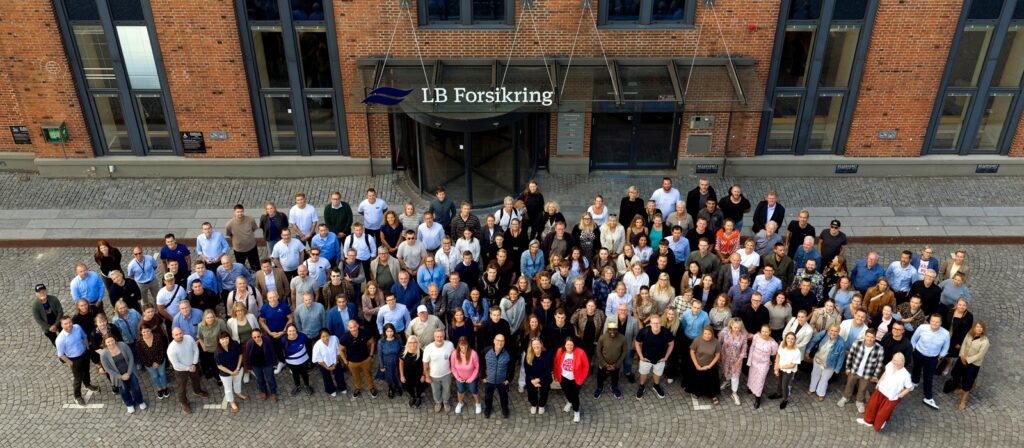 A large group of people stands together outside a brick building with the LB Forsikring sign above the entrance, posing for a group photo on a cobbled pavement.
