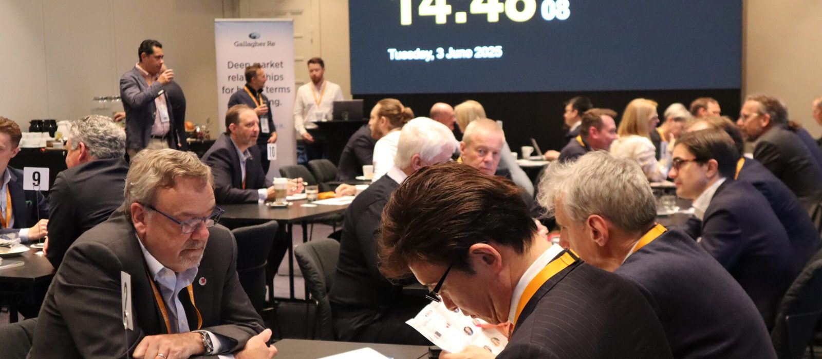 A group of people in business attire sit at round tables in a conference room, networking and discussing. A large digital clock and date are displayed on a screen in the background, reading 14:46:08, Tuesday, 3 June 2025.