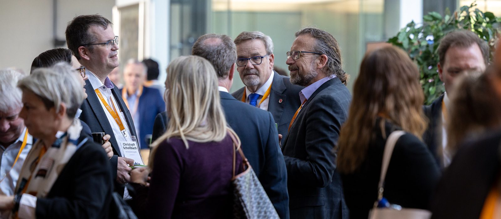 A group of people, mostly in business attire with orange lanyards, are gathered indoors, engaged in conversation at what appears to be a professional networking event or conference.