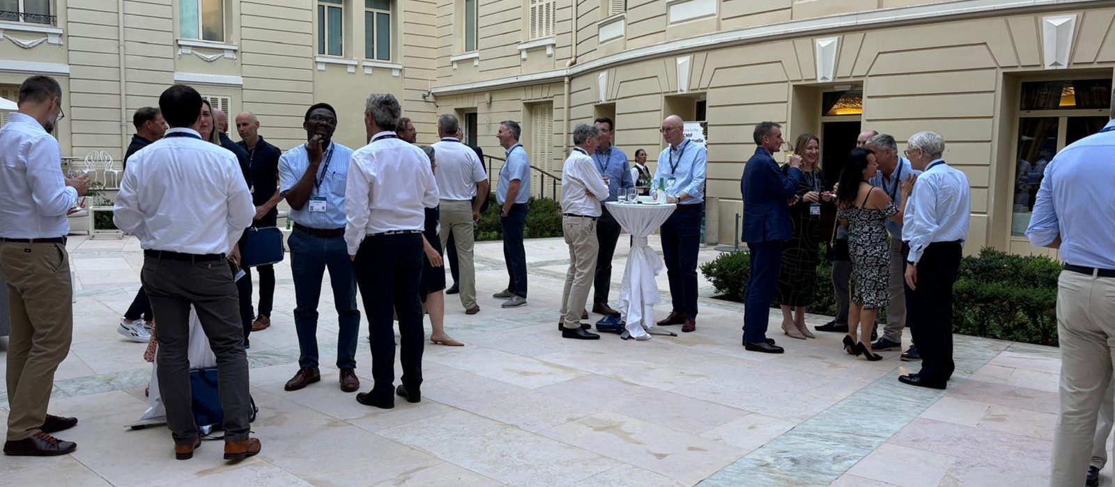 People dressed in business or smart casual attire are mingling and talking in an outdoor courtyard. Small groups are gathered around tall tables with drinks, and a beige building with windows is in the background.