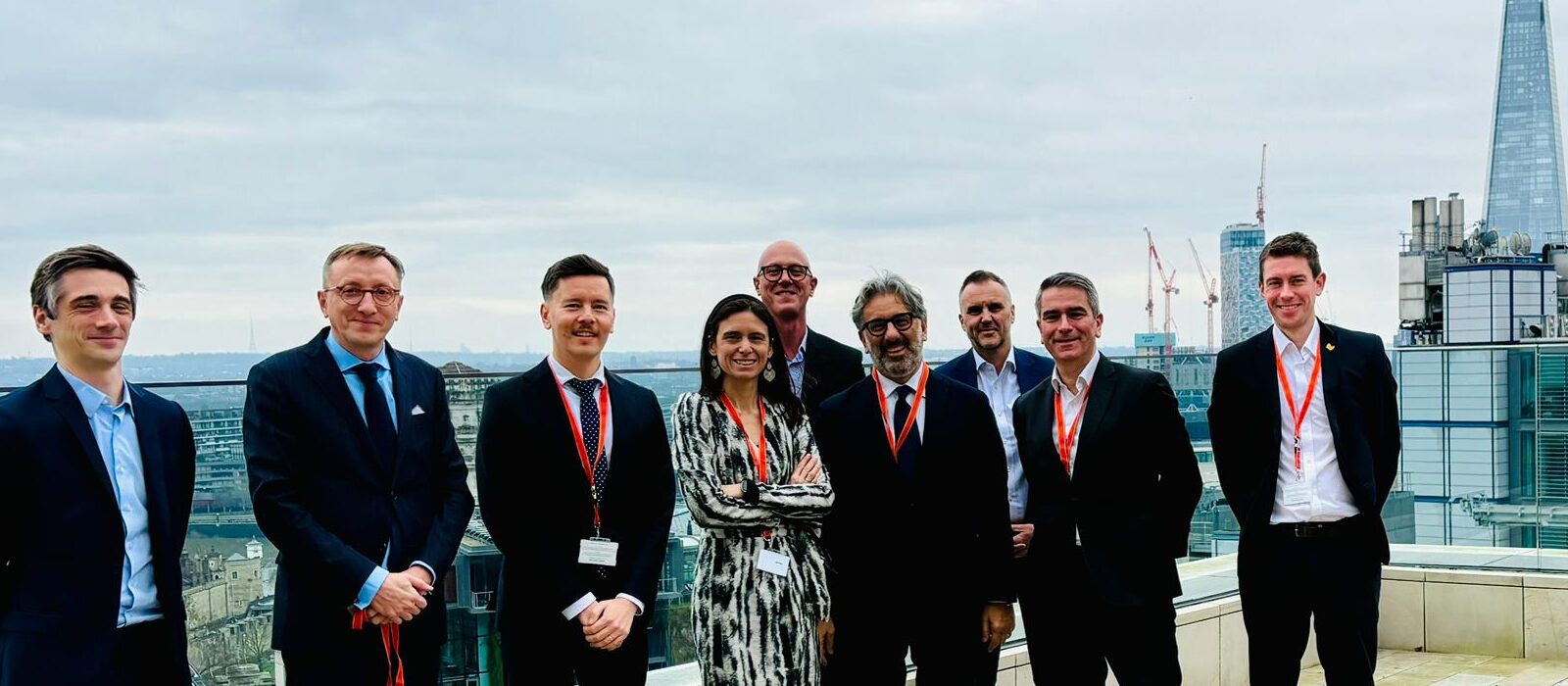 A group of nine people in business attire, wearing red lanyards, stand on a rooftop with a city skyline and tall buildings, including the Shard, in the background under a cloudy sky.