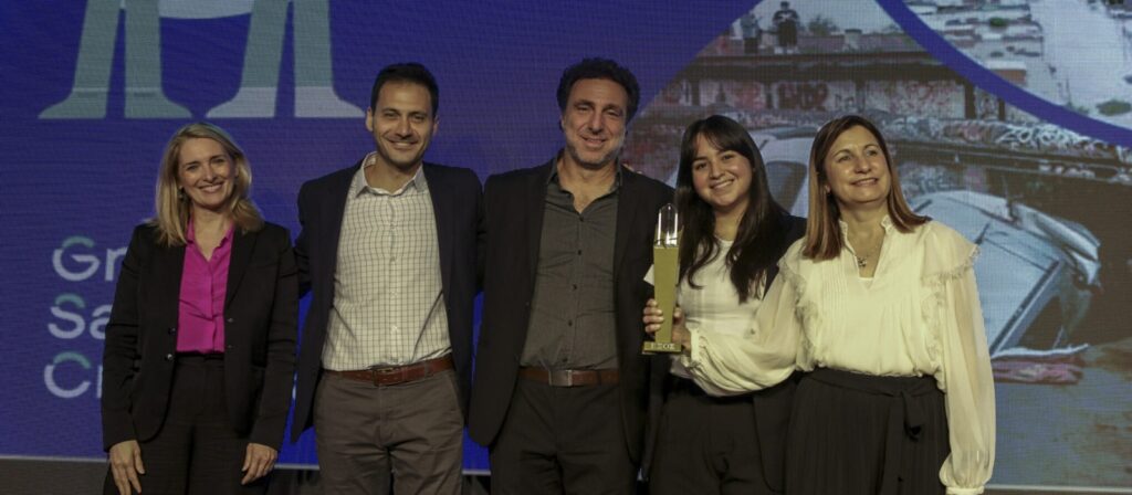 Five people in business attire stand together on stage, smiling at the camera. One woman in the centre holds a trophy. A large screen with graphics and text is visible in the background.
