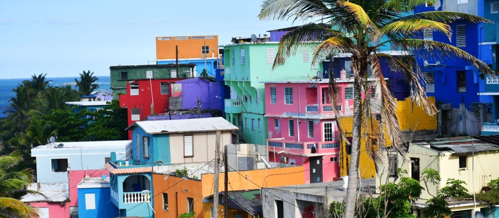 Colourful buildings in shades of blue, red, yellow, green, and pink are stacked on a hillside near the ocean, surrounded by palm trees under a clear blue sky.