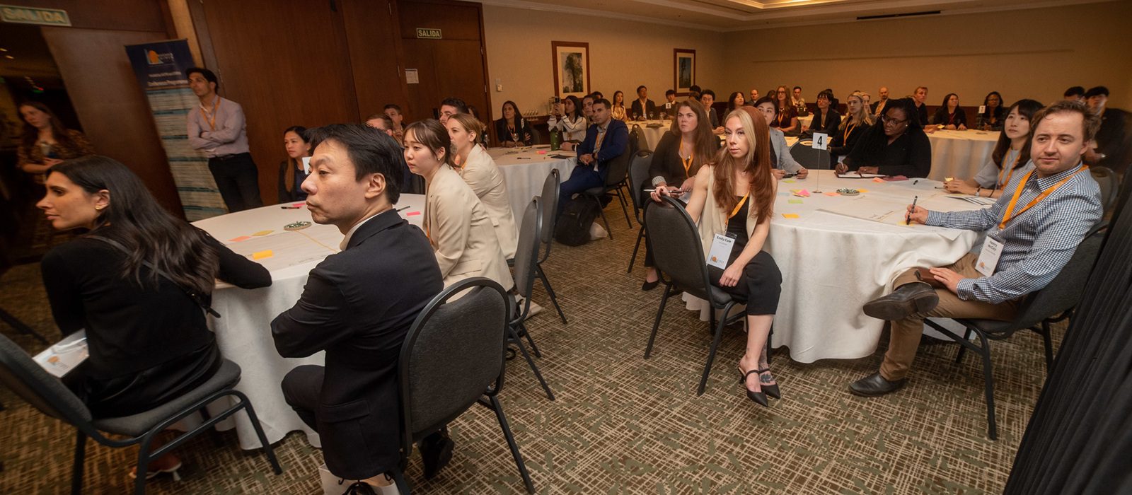 A group of people sits around round tables in a conference room, attentively listening to a presentation. Most are wearing business attire and name badges. Some are taking notes; the atmosphere appears professional and engaged.