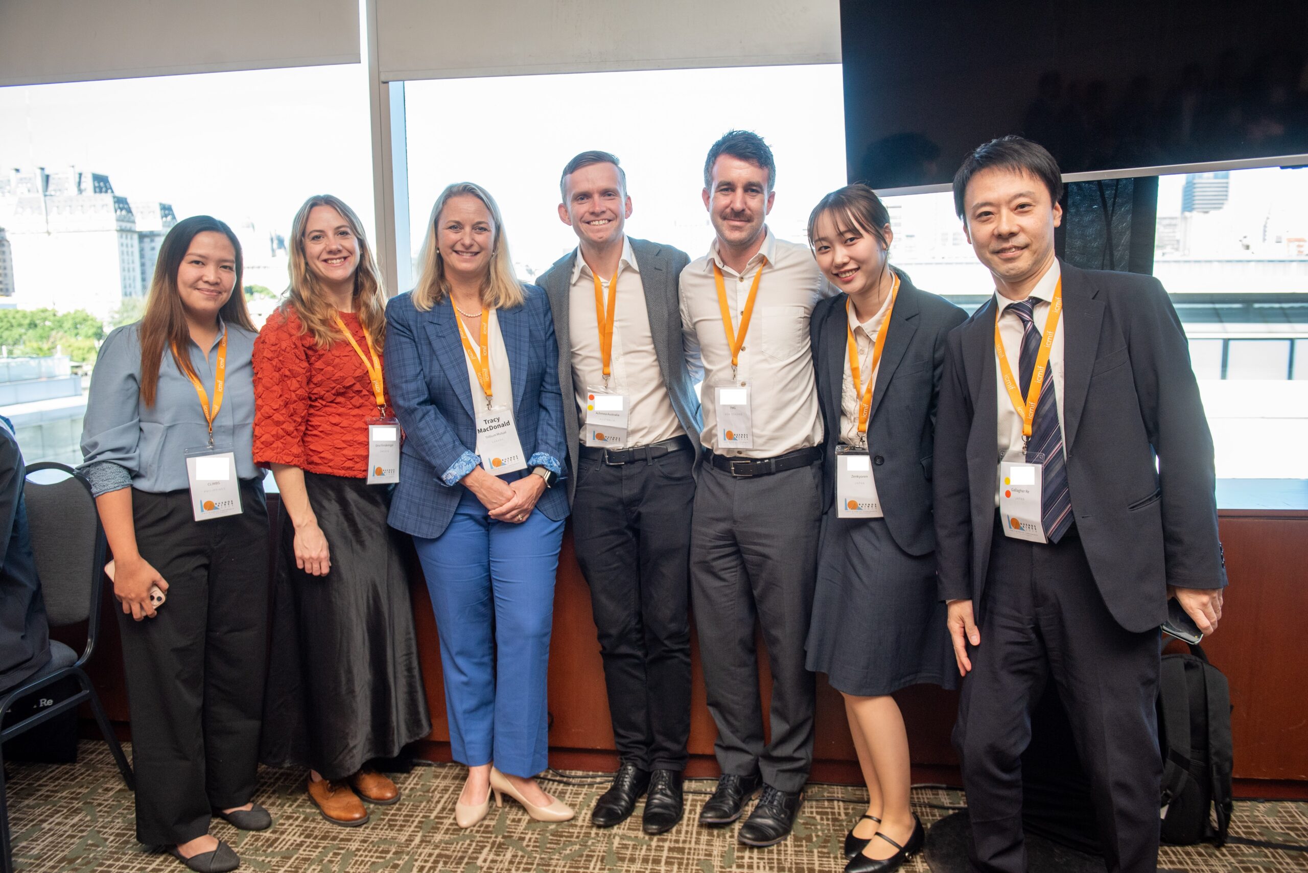 Seven people wearing business attire with orange lanyards stand together indoors, smiling at the camera. Large windows behind them show an urban cityscape in daylight.
