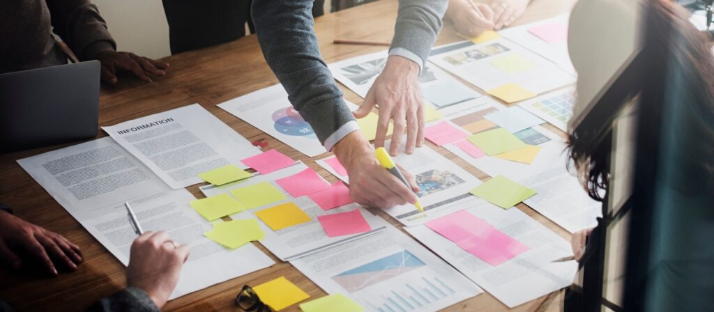A group of people work together at a table covered with documents, charts, and colourful sticky notes. One person highlights a paper while others discuss, suggesting a collaborative planning or brainstorming session.