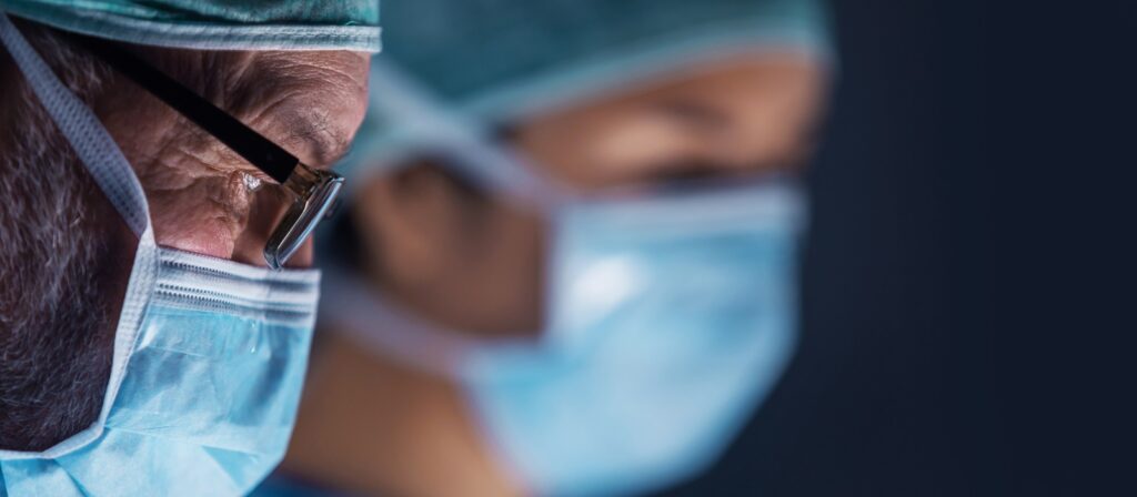 Close-up of two healthcare professionals wearing surgical masks, hair covers, and glasses, focused on a task in a medical setting. The background is dark and blurred, highlighting their concentration.
