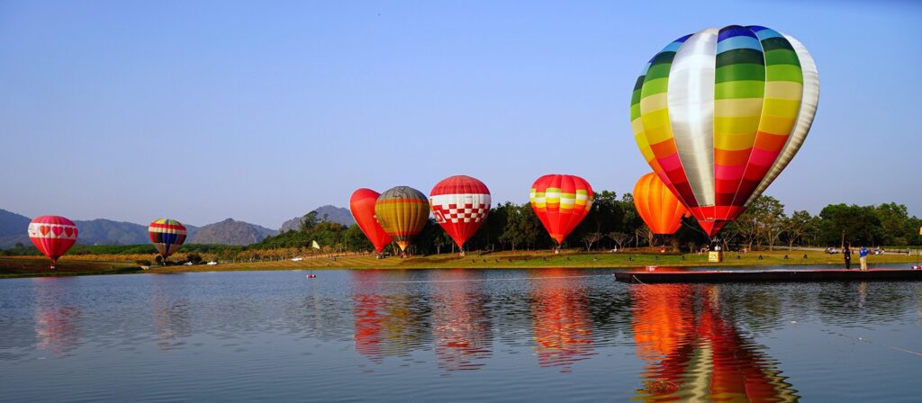 Colourful hot air balloons float above a calm lake, reflecting on the water’s surface. Green hills and trees line the background under a clear blue sky.