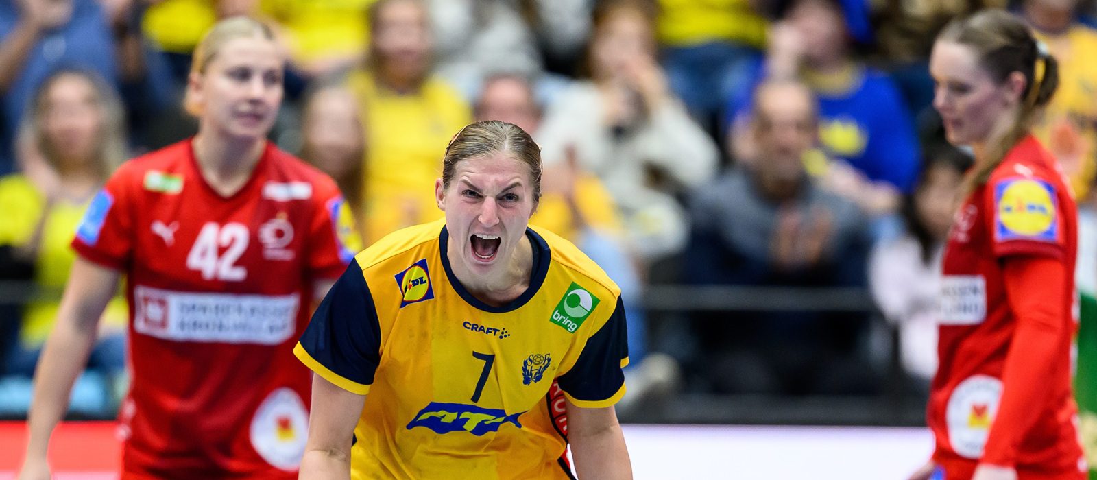 A handball player in a yellow and blue kit shouts energetically on the court, flanked by two players in red kits. The background shows a blurred crowd wearing yellow.