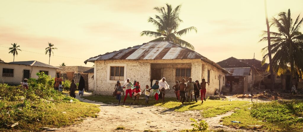 Children run and play outside a rustic house with a rusted metal roof, surrounded by palm trees and greenery, under a warm, golden sky. Other houses are visible in the background.