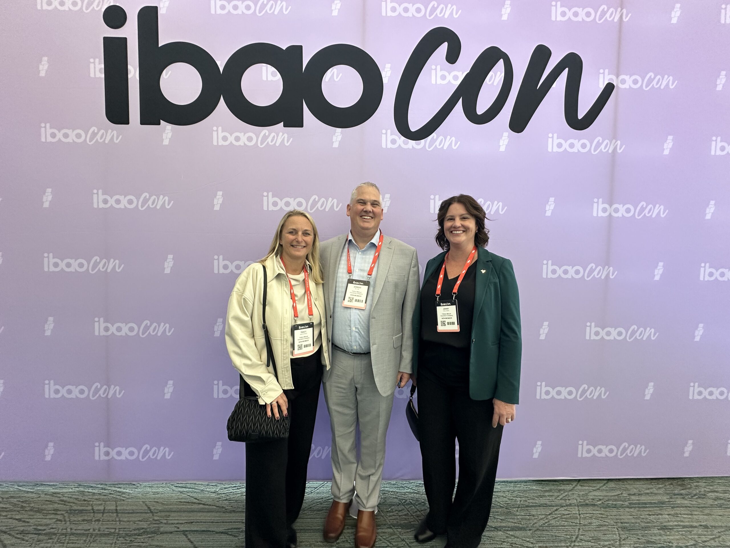 Three people wearing conference badges stand smiling in front of a purple backdrop with the repeated logo ibao con. Two women are on either side of a man dressed in a light grey suit.