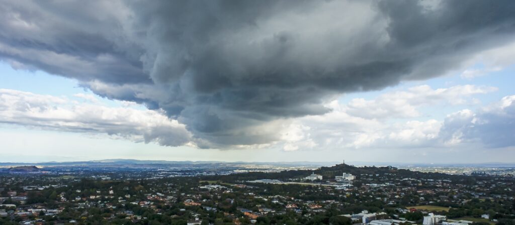 A large, dark storm cloud hangs low over a cityscape, casting a shadow on the buildings and landscape below, with patches of blue sky and distant hills in the background.