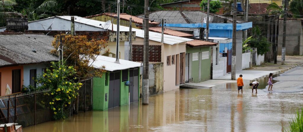 Flooded residential street with muddy water. Three children stand and play in the water near colourful houses. Vegetation and utility poles are visible along the road. The scene appears calm despite the flooding.