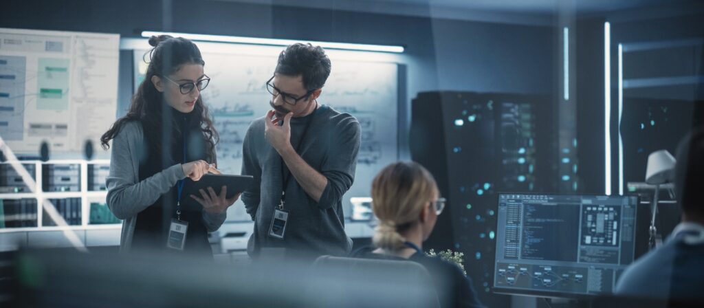 Two people in a modern office look at a tablet and discuss information. Computer monitors, technical diagrams, and servers are visible, creating a high-tech, collaborative work environment.