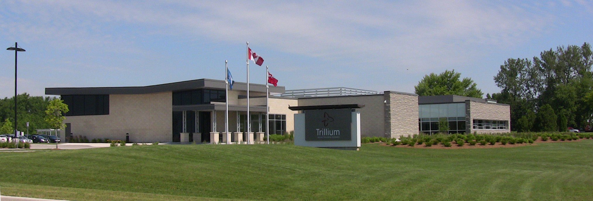Modern, single-storey building with large windows and a Trillium sign in front, three flagpoles flying flags, surrounded by green lawns and trees under a partly cloudy sky.