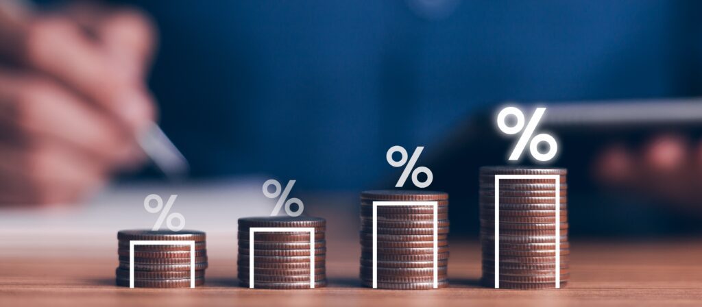 Stacks of coins arranged in ascending order with percentage signs above each stack, representing increasing savings or financial growth. A blurred person holding a pen and device is visible in the background.