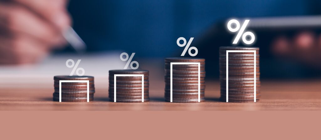 Stacks of coins arranged in ascending order with percentage symbols above each stack, symbolising increasing interest or financial growth, with a blurred hand and tablet in the background.