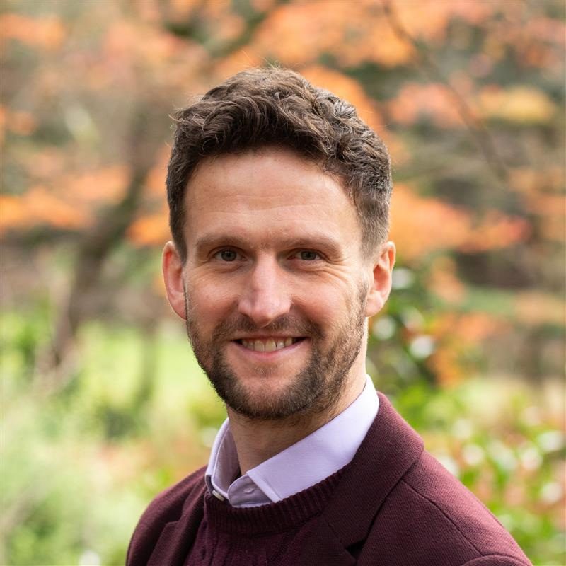A man with short brown hair and a beard smiles outdoors, wearing a maroon jumper over a collared shirt. The background features blurred autumn foliage in orange and green hues.