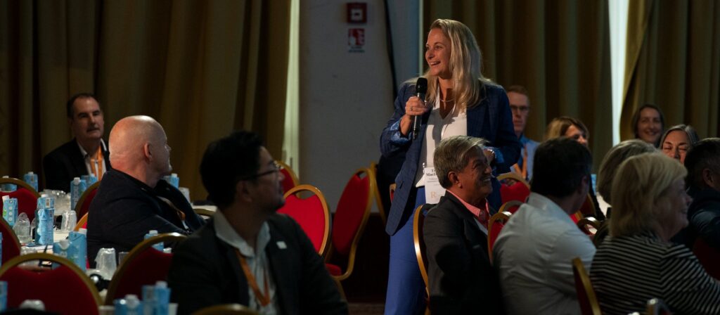 A woman stands and speaks into a microphone during a conference, smiling, surrounded by seated attendees at round tables with water bottles. The room has red chairs and curtains.