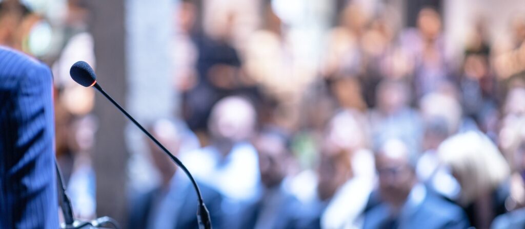 A close-up of a microphone on a stand in the foreground, with a blurred audience seated and watching in the background, suggesting a conference or public speaking event.