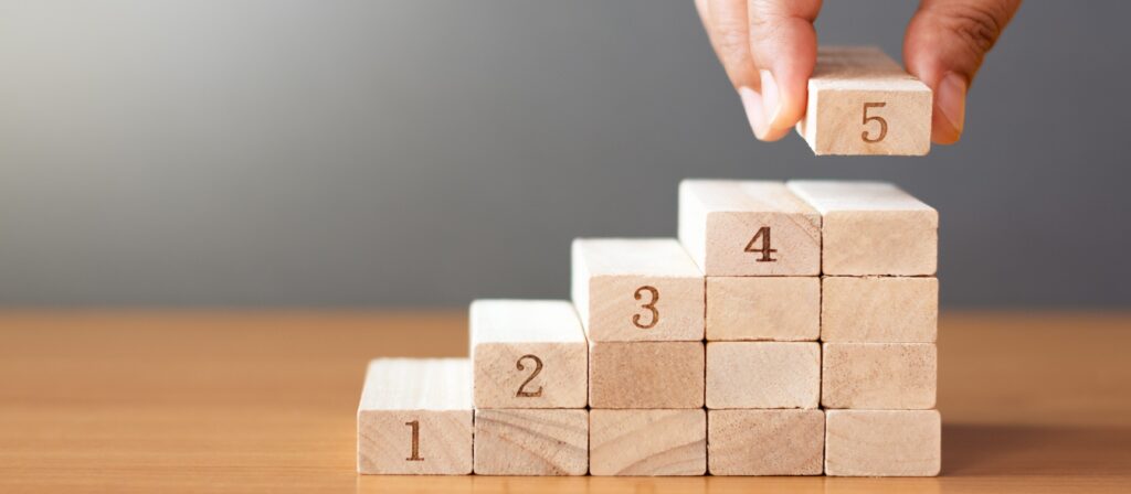 A hand places a wooden block labelled 5 on top of a staircase made of wooden blocks numbered 1 to 4, arranged in ascending order on a wooden surface with a plain background.