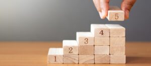 A hand places a wooden block labelled 5 on top of a staircase made of wooden blocks numbered 1 to 4, arranged in ascending order on a wooden surface with a plain background.