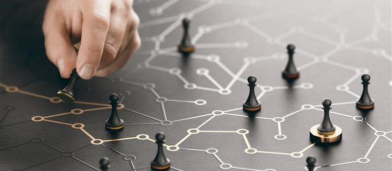A hand moves a chess pawn on a board featuring interconnected lines and nodes, symbolising network connections and strategic planning. Other pawns are placed at various points along the network.