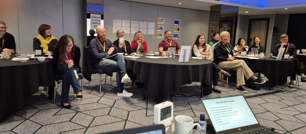 A group of people sit in a conference room, smiling and laughing during a meeting. Round tables with papers, drinks, and laptops are visible. A clock, water bottle, and presentation slides are seen in the foreground.