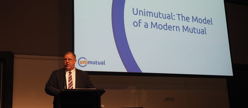 A man in a suit speaks at a lectern in front of a screen displaying the text Unimutual: The Model of a Modern Mutual and the Unimutual logo.