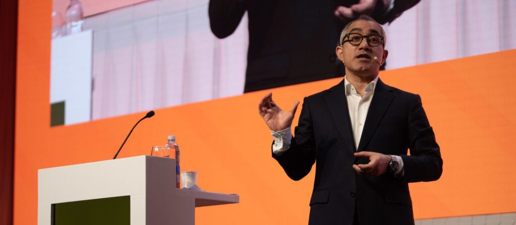 A man in a suit speaks on stage with a headset microphone, gesturing with one hand. A lectern with a water bottle and cup is nearby. A large screen behind him displays his image on an orange background.