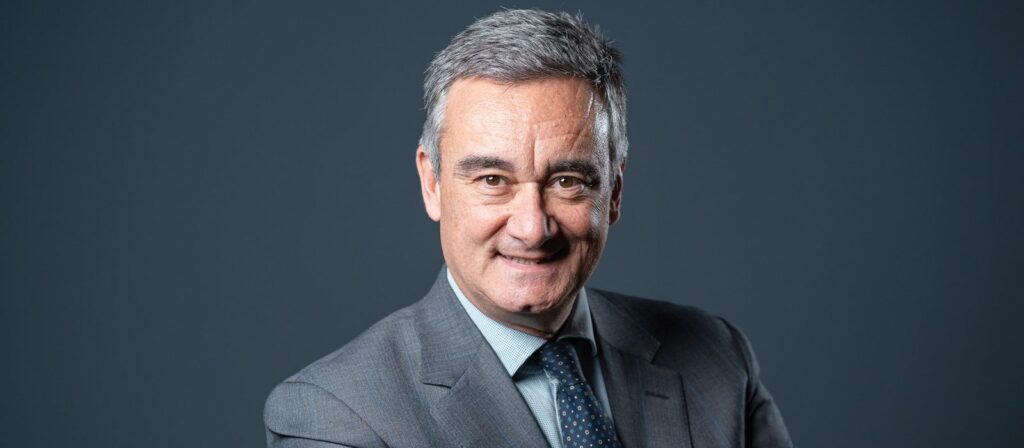 A middle-aged man with grey hair wearing a grey suit, blue shirt, and polka dot tie, smiling and standing against a plain dark background.