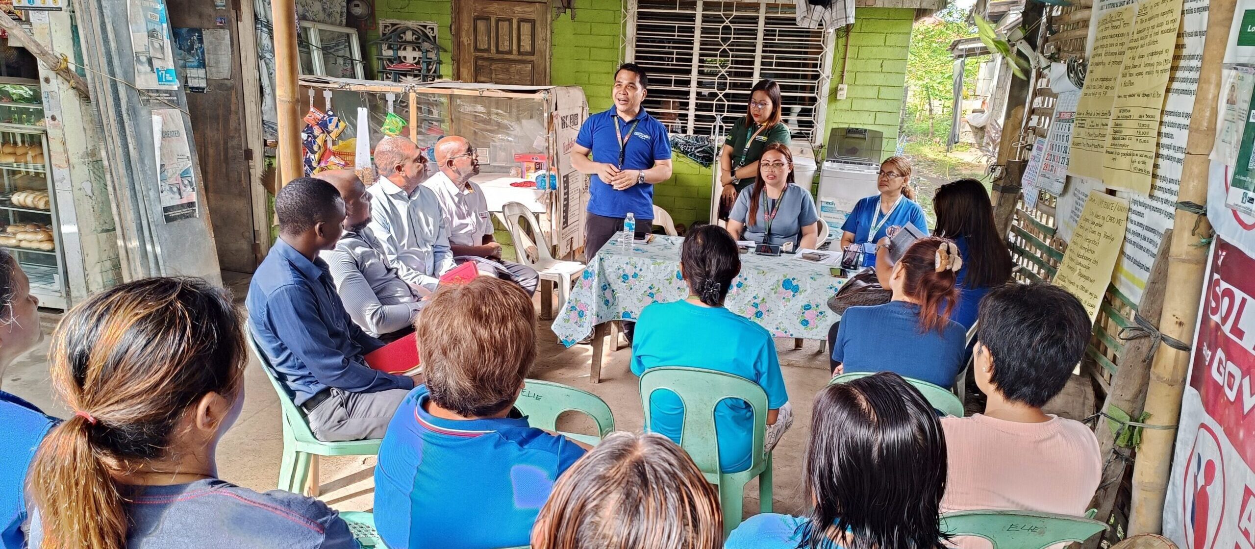 A group of people sit in plastic chairs facing a table with several speakers in front of a small shop, attentively listening to a man standing and speaking during a community meeting.