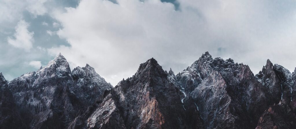 Jagged mountain peaks with rocky, dark surfaces are dusted with snow under a cloudy sky, creating a dramatic and rugged landscape scene.
