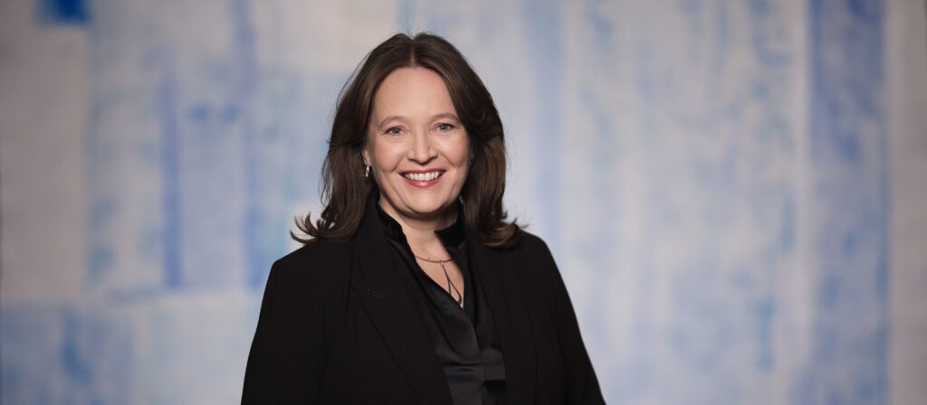 A woman with long brown hair, wearing a black blazer and blouse, smiles whilst standing in front of a blue and white abstract background.
